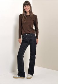 Braunes Langarmoberteil, dunkelblaue Jeans mit Kontrastnähten und weiße Sneaker mit hellblauen Akzenten. Das Modell steht vor einem neutralen Hintergrund.