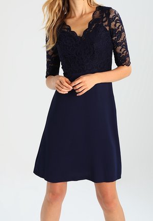 Kvinde iført en mørkeblå kjole med blomsterblonde korte ærmer og korset samt en ensfarvet A-formet nederdel, stående med hænderne foldede.