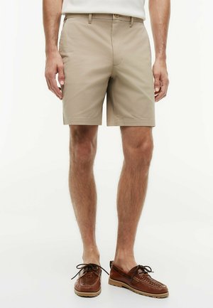Mand iført beige skræddersyede shorts og brune læder snørestøvler, stående foran en ensfarvet hvid baggrund.