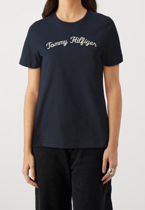 Osoba nosząca granatową koszulkę z krótkim rękawem z wyhaftowanym na piersi napisem "Tommy Hilfiger" oraz czarne spodnie.