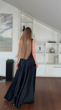 Schwarzes bodenlanges Kleid aus glattem Stoff mit einem fließenden Rock und ärmellosem Design. Der Hintergrund zeigt minimalistische Regale.