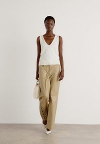 Débardeur blanc en tricot à col en V avec un ourlet côtelé, associé à un pantalon en simili cuir beige clair, ajusté avec des lacets sur les côtés, et des chaussures blanches à talons pointus.