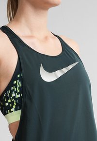 Donker groene sportieve tanktop met een ronde halslijn. Heeft een groot zilver Nike-logo en patroon details in tinten groen en geel aan de zijkant.