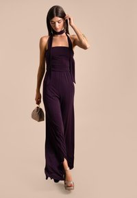 Mono morado con parte superior sin tirantes, pantalones fluidos y aberturas laterales; combinado con un bolso gris y zapatos de plataforma. Textura de tela suave y lisa.