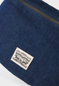 Jeansov pouches v temno modri tkanini z zlatim zadrgo. Vključuje pravokotno bež oznako s črnim tiskom logotipa Levi Strauss & Co.