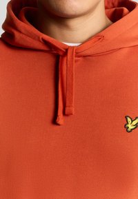 Sudadera naranja con cordón en el cuello y un pequeño logo bordado de un pájaro amarillo en el lado izquierdo del pecho. Textura de tela suave.