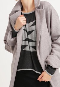 Übergroßer grauer Mantel mit weitem Kragen, über einem geometrisch gemusterten Pullover in Schwarz und Grau geschichtet. Weiche, strukturierte Stoffe mit kontrastierenden Besatz.