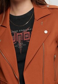 Rdzawy, teksturowany sweter z srebrnymi detalami, zamykanymi kieszeniami i kołnierzem klapowym, nałożony na czarny t-shirt z grafiką.