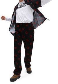 Pantalon noir avec motif floral rouge, assorti à une chemise blanche à manches longues et une veste foncée, porté avec des chaussures marron.