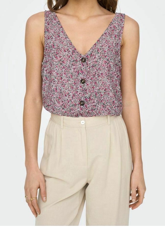 Femme portant un chemisier sans manches rose à motifs floraux boutonné, rentré dans un pantalon beige plissé taille haute, avec des bagues aux doigts.