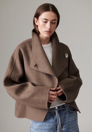 Femme portant un manteau en laine marron oversize avec de larges manches et un grand col par-dessus une chemise blanche et un jean bleu sur un fond uni.