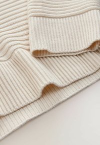 Krémová ribbovaná pletenina přeložená, aby ukázala texturu a tloušťku, zvýrazňující detaily měkkého, útulného materiálu.