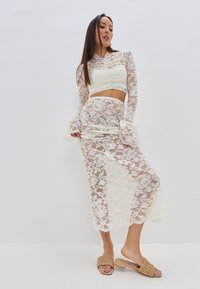 Set composto da top corto in pizzo e gonna color avorio, con motivi floreali, maniche lunghe in pizzo e polsini a campana con rouches, abbinato a sandali beige scivolati.