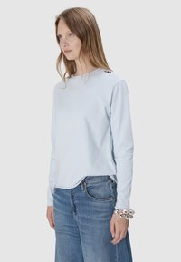 Hellblaues Langarmhemd aus weichem Stoff mit rundem Ausschnitt und Knopfdetail an der Schulter, kombiniert mit blauen Jeans.