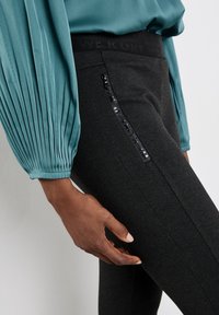 Svarta leggings med texturerad yta, med en zipficka på sidan och en svart märkesmidja. Följt av en teal-blus.