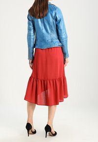 Frau trägt eine blaue Lederjacke, einen roten Midirock mit Rüschenbesatz und schwarze High Heels, von hinten vor einem weißen Hintergrund gezeigt.