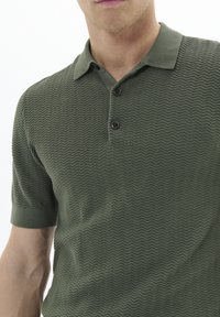 Cămașă polo verde cu o textură ondulată, având un guler scurt și trei nasturi negri la decolteu. Design slim fit cu mâneci scurte.