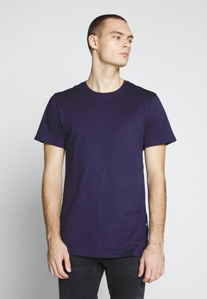 Camiseta de manga corta azul marino hecha de algodón suave, con cuello redondo y pequeño detalle de logo en el pecho, combinada con jeans oscuros.