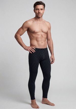 Hemdloser Mann mit kurzen dunklen Haaren, der schwarze gemusterte lange Unterwäsche trägt, steht barfuß vor einem grauen Hintergrund.