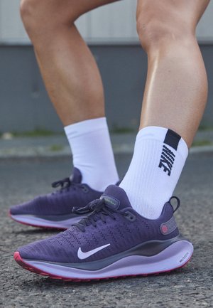 Paarse Nike-hardloopschoenen met een gestructureerd bovenwerk, witte Swoosh, roze accenten op de zool, en gecombineerd met witte sportsokken.