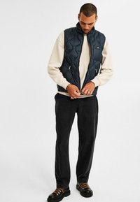 Man i beige sweatshirt, marinblå vadderad väst, svarta byxor och svarta stövlar som drar upp västen, stående mot vit bakgrund.