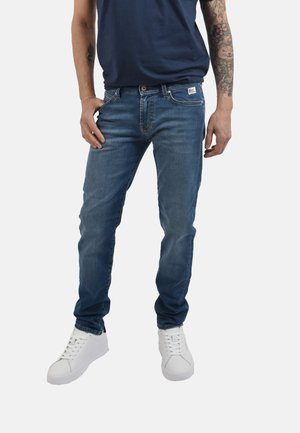 Uomo che indossa jeans blu slim fit, t-shirt navy, sneakers bianche, con il braccio destro tatuato e la mano sinistra in tasca.