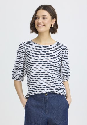 Femme aux cheveux courts bruns portant des boucles d'oreilles en or, un haut blanc à rayures ondulées noires et un pantalon bleu taille haute avec les mains dans les poches.