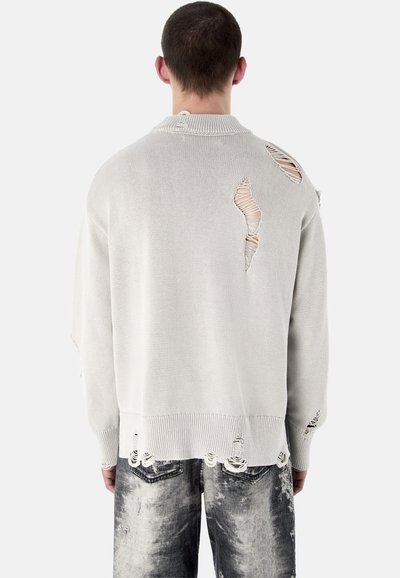 Osoba nosząca jasnoszary sweter z przetarciami z dużymi rozdarciami na plecach i rękawach, w połączeniu z wyblakłymi czarno-białymi dżinsami.