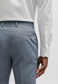 Grå texturerade byxor med slim fit, matchade med en vit skjorta med knäppningsmanschetter. Detalj med bakficka synlig.