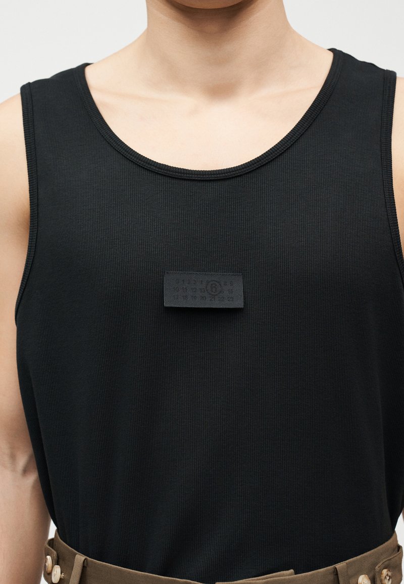 Primer plano de una persona vistiendo una camisa negra sin mangas con un pequeño parche negro en el centro del pecho, metida dentro de unos pantalones marrones con botones.