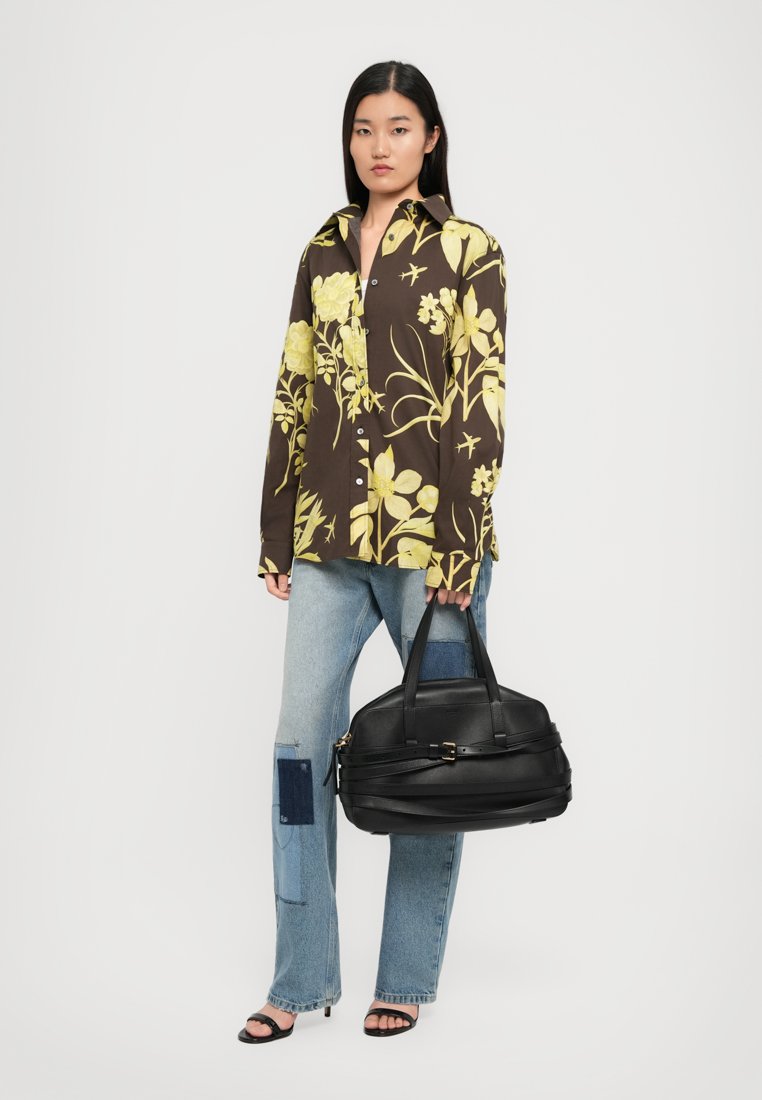 Sac à main en cuir noir avec doubles poignées, texture douce et subtilement décoré de quincaillerie. Le modèle porte une chemise à motifs floraux et un jean clair délavé.