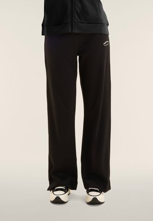 Persona che indossa pantaloni neri a gamba larga con scritta bianca sulla coscia e sneakers bianche e nere, in piedi davanti a uno sfondo semplice.