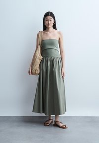 Γυναίκα που φορεί ελιάς πράσινο strapless μάξι φόρεμα, κρατώντας τσάντα ώμου από ψάθα σε ταμπά χρώμα και καφέ σανδάλια, στέκεται μπροστά σε ανοιχτογκρι τοίχο.