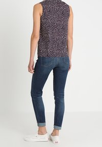 Osoba stojí zády, má na sobě bezruko se černým topem s bílými tečkami, roztrhané tmavě modré džíny a bílé slip-on tenisky.