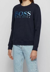 En person som bär en marinblå tröja med lång ärm med "BOSS HUGO BOSS" i blåa gradientbokstäver och ljusblå denimjeans.