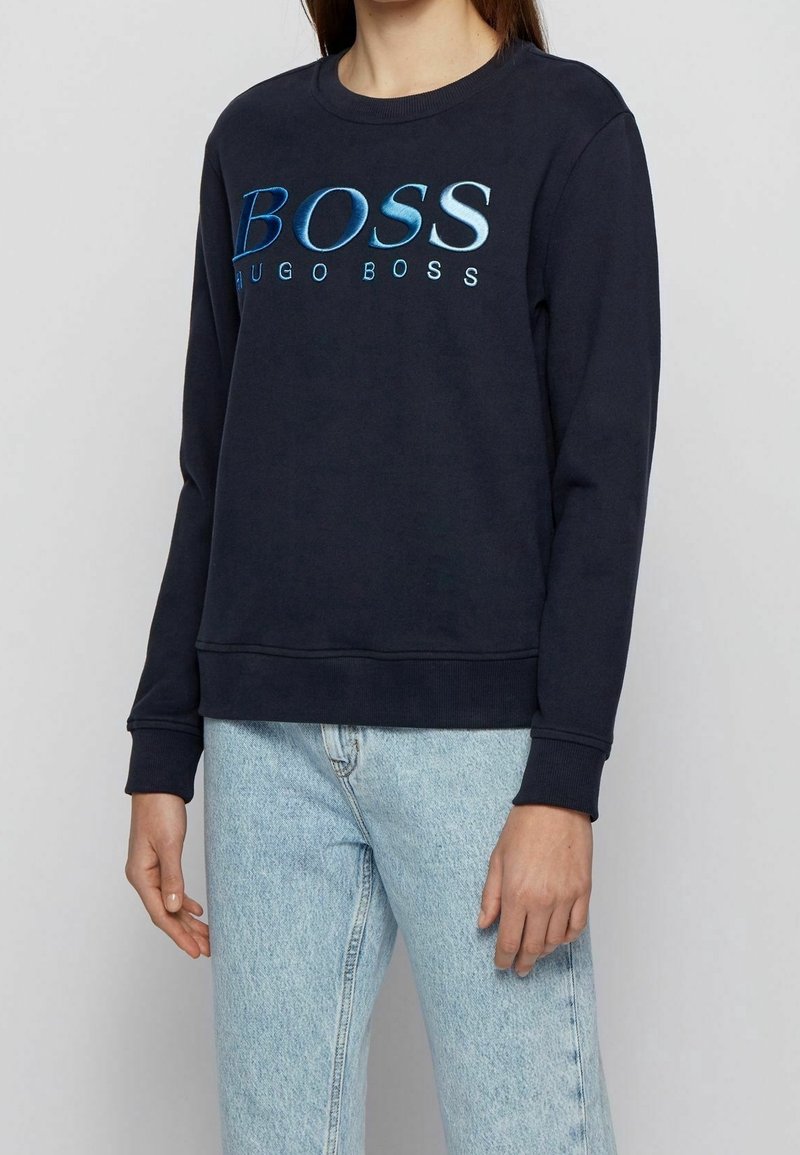 En person som bär en marinblå tröja med lång ärm med "BOSS HUGO BOSS" i blåa gradientbokstäver och ljusblå denimjeans.