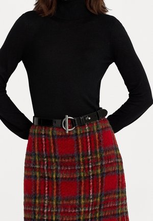 Frau trägt einen schwarzen Rollkragenpullover mit langen Ärmeln, der in einen rot-grün-gelb karierten Rock gesteckt ist, dazu einen schwarzen Gürtel mit silberner Schnalle.