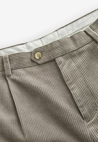 Beige corduroy trousers with vertical ribs, featuring a front fold and a single brass button at the waistband. White inner lining visible.