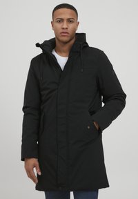 Abrigo parka negro con capucha, con cierre de cremallera, bolsillos laterales y cordones ajustables. Corte ajustado y textura suave.