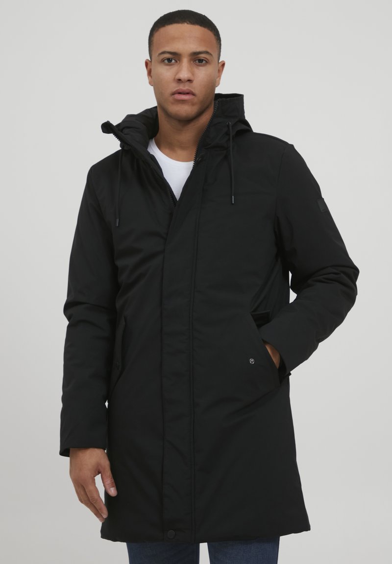 Abrigo parka negro con capucha, con cierre de cremallera, bolsillos laterales y cordones ajustables. Corte ajustado y textura suave.