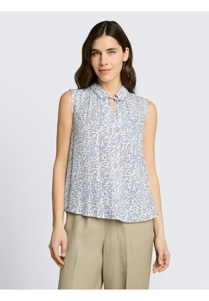 Kobieta ubrana w białą, bezrękawną bluzkę z niebieskim kwiatowym wzorem i beżowe spodnie o szerokich nogawkach, stojąca na tle jasnoszarego tła.