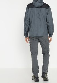 Harmaa vedenpitävä takki mustilla yksityiskohdilla; väljä malli, elastiset hihansuut ja säädettävä huppu, yhdistettynä harmaisiin cargo-housuihin ja tummiin jalkineisiin.