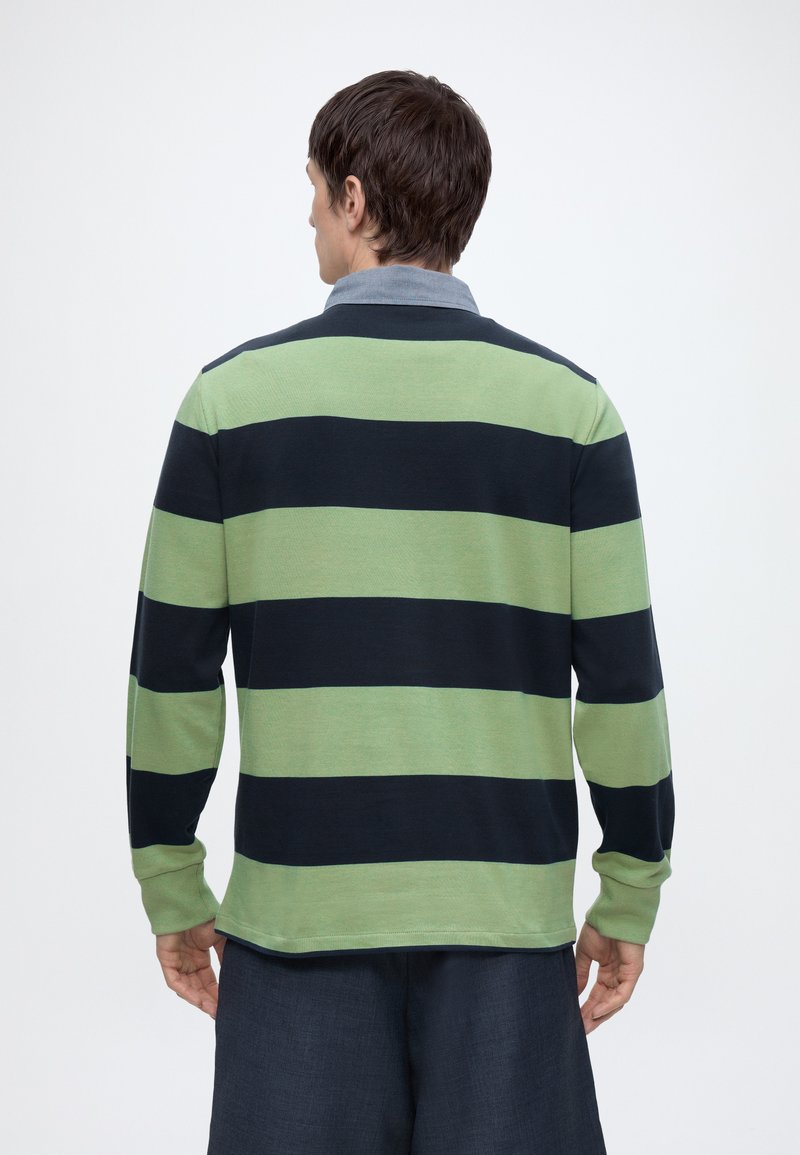 Personne de dos, portant un maillot de rugby à manches longues rayé horizontalement vert et noir et un pantalon sombre, sur un fond uni.