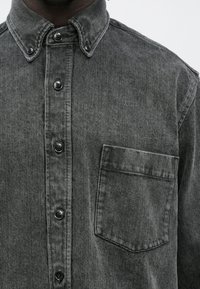 Camisa de ganga cinzenta com gola abotoada, bolso frontal e botões pretos. Apresenta um tecido texturado e um ajuste descontraído.