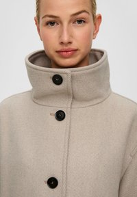 Beige wollen jas met een hoge kraag en drie zwarte knopen. Heeft een gestructureerde afwerking en een recht model. Minimalistische styling.