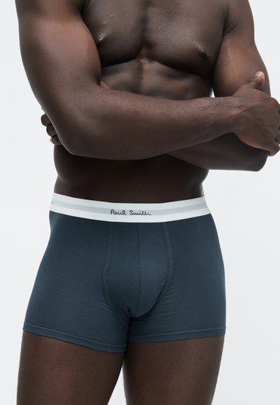 Homme musclé à la peau foncée, torse nu, portant un boxer bleu foncé avec une ceinture blanche estampillée Paul Smith, bras croisés sur la poitrine.
