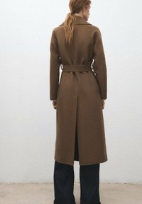 Brązowy płaszcz z paskiem, kołnierzem, rozcięciem z tyłu i długimi rękawami, prezentowany z ciemnymi spodniami. Tkanina wydaje się gładka i dobrze uformowana.