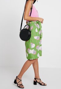 Jupe verte à motifs avec des designs floraux blancs et à pois, associée à un haut rose et un sac bandoulière noir en forme de cercle. Sandales noires à lanières.