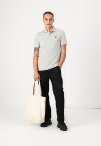 Světle šedý polo svetr s malým žlutým logem na hrudi, doplněný černými kalhotami. Model drží béžovou plátěnou tašku.