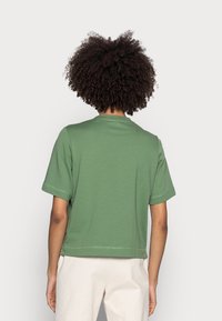 Persona con capelli ricci che indossa una maglietta verde a maniche corte e pantaloni color avorio, vista di spalle su uno sfondo semplice.